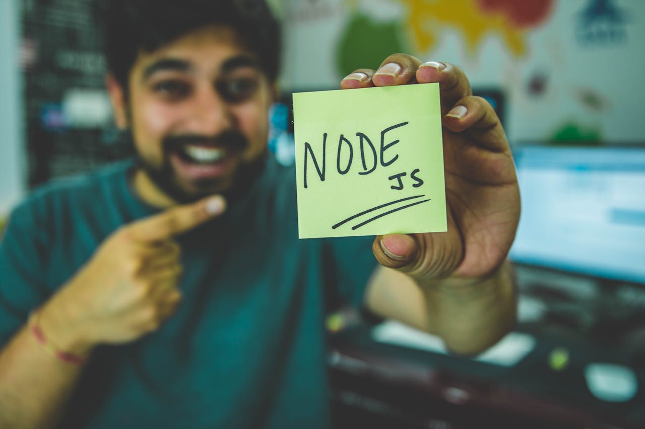 Home A cheerful software developer shows a Node.js sticky note in a tech workspace.